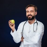 Portrait of a happy male doctor dressed in uniform with stethoscope holding green apple and looking at camera isolated over dark background